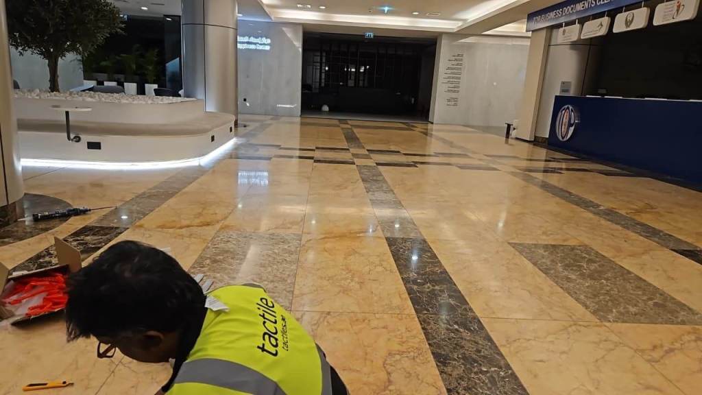 An employee in a high-visibility vest is seen kneeling on a polished marble floor in a well-lit lobby area. There is modern seating and a decorative planter in the background, along with a reception desk displaying signage.