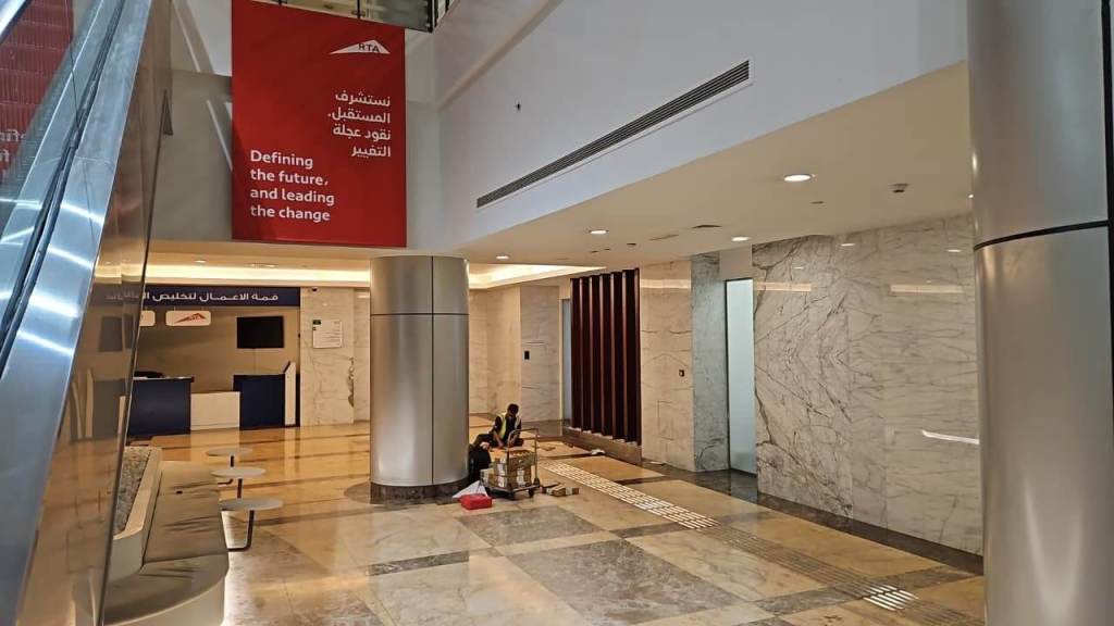 Interior view of a modern building with marble flooring and walls, featuring a sign that reads 'Defining the future, and leading the change.' A reception area in the background with blue accents and a staff member present.