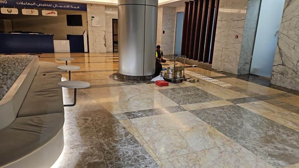A person wearing a yellow vest sits on the floor near a cart with boxes in a spacious, marble-floored lobby area, featuring modern seating and a reception desk in the background.