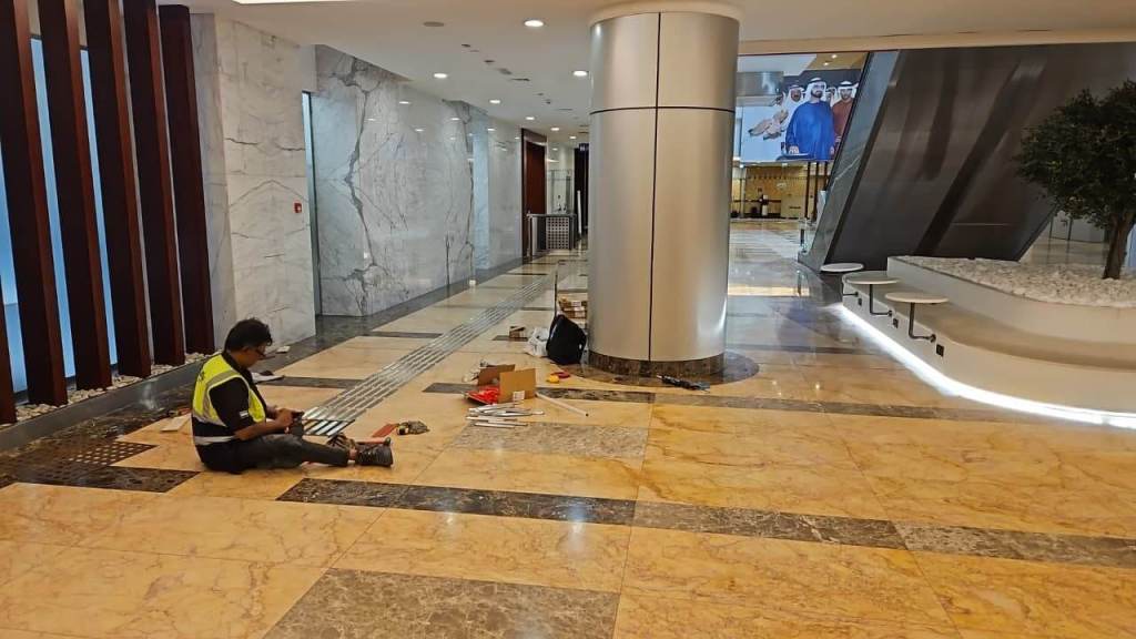 A construction worker sitting on the floor of a modern lobby, wearing a safety vest, surrounded by tools and materials on the polished stone floor.
