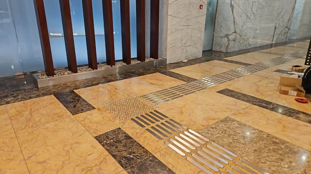 Interior view of a modern building lobby featuring polished marble flooring with metal strips and tactile indicators for navigation, alongside a decorative wooden feature wall and potted stones.