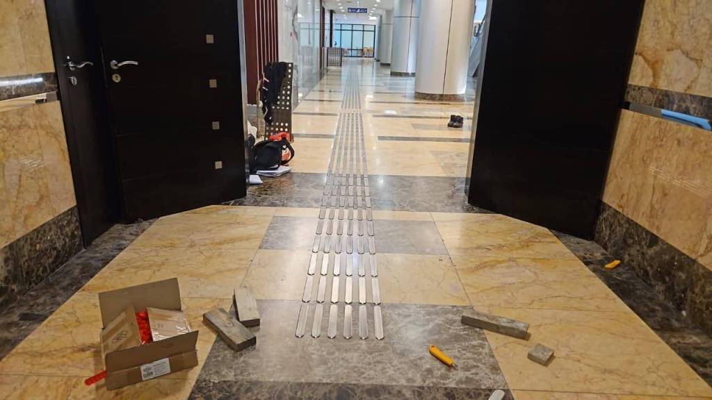 Interior view of a hallway with dark doors and a tactile guide strip in the center, surrounded by scattered construction materials like bricks and tools.
