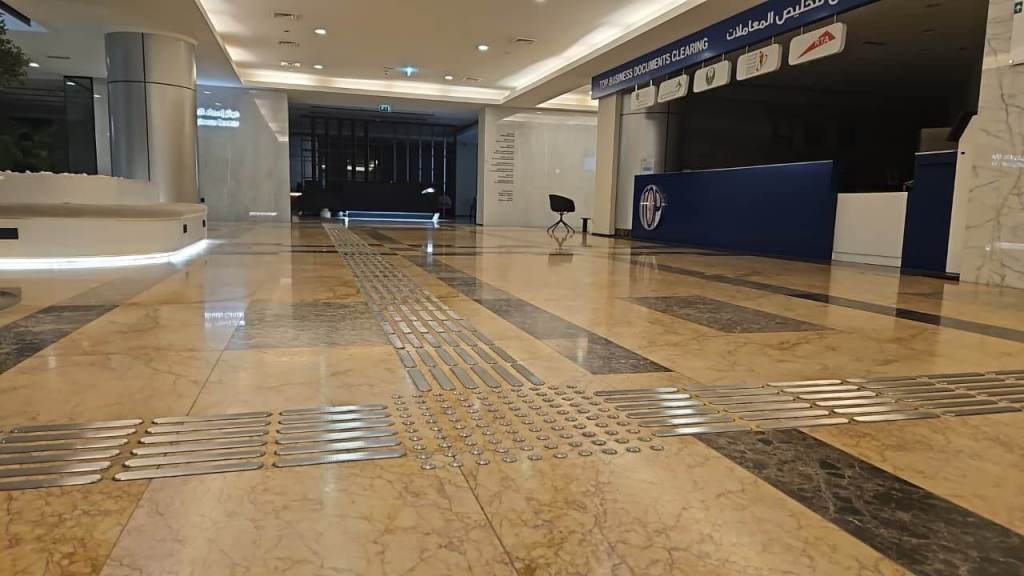 Interior view of a modern commercial lobby with polished marble flooring, featuring tactile guide strips for accessibility and a reception area in the background.