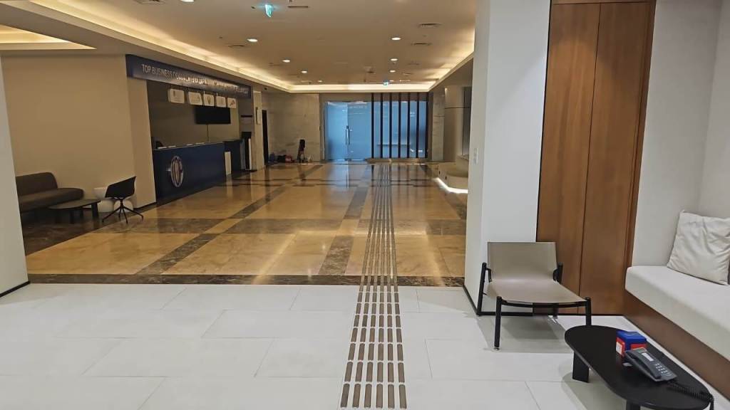 A modern lobby with a reception desk, seating areas, and a marble floor. The space features a pathway indicator and glass doors at the far end.