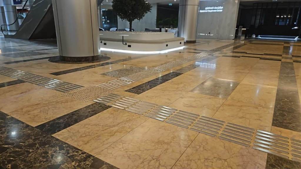 A close-up view of a marble floor featuring shiny metal tactile strips, with a fountain and greenery visible in the background.
