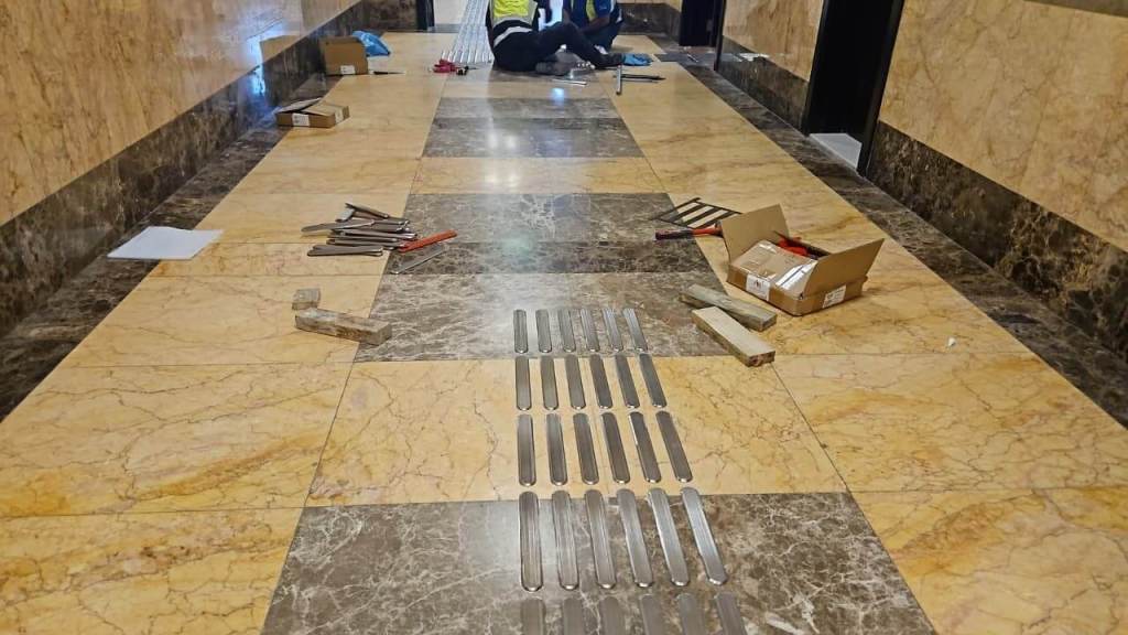 A construction area in a hallway featuring marble tiles with installation materials scattered around, including metal strips, tools, and a box.