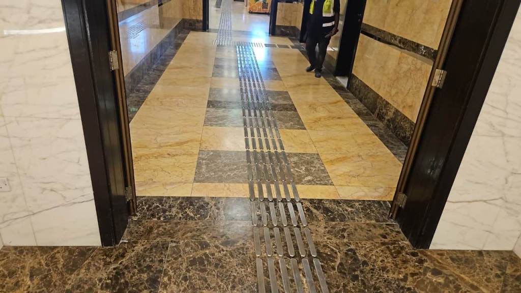 Interior hallway with polished marble flooring and metallic drainage strips, featuring a person walking in a safety vest.