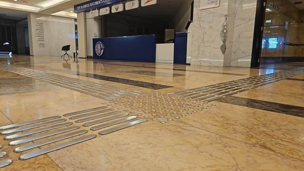 Interior view of a spacious lobby featuring textured floor tiles and tactile ground surface indicators. In the background, a reception desk is visible with signage.