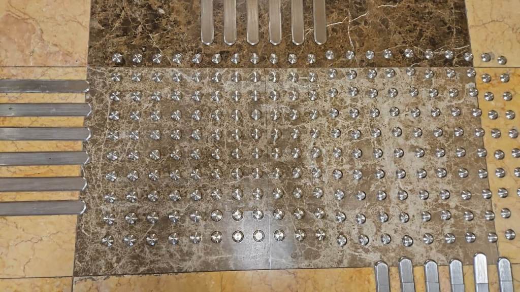 A close-up view of a textured floor surface featuring arranged metal strips and circular studs, possibly designed for slip resistance or tactile guidance.