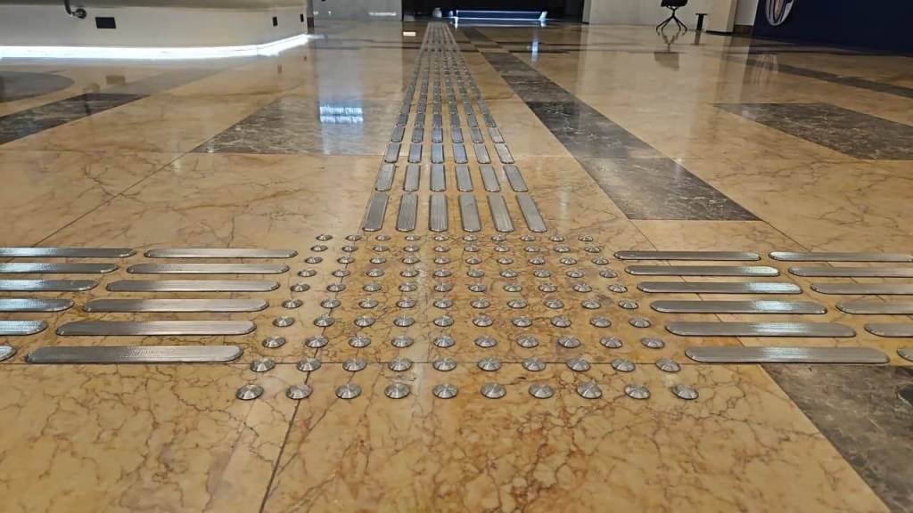 Close-up view of a textured floor surface with metal tactile indicators arranged in lines and patterns, designed for accessibility.
