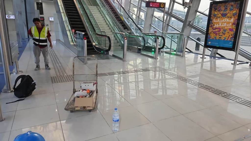 An indoor transportation hub featuring escalators, a man in a safety vest, a cart with tools, a backpack, and a water bottle on the polished floor.