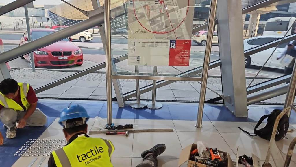 Construction workers installing tactile paving in a building entrance, with a map displayed nearby and a red car visible outside.