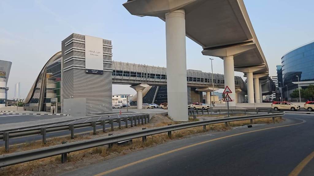 A modern urban scene featuring a curved building and an elevated structure for transportation, surrounded by a roadway and low vegetation. Several vehicles are visible on the road.