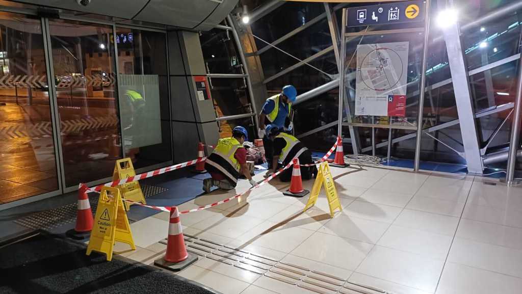 Workers in safety vests performing maintenance in a transit station at night, with caution signs and construction barriers around the area.