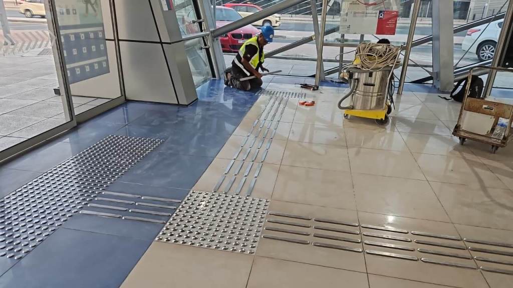 A worker installing tactile paving and floor tiles in a large indoor space with glass walls and metal supports.