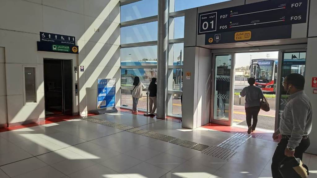 Interior of a modern transport station featuring large windows, an elevator, and informational signage. People are walking towards the exit, with a bus visible outside.
