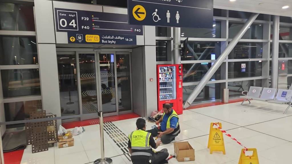 Interior of an airport with Gate 04 signage, showing gates F39 and F40. Two workers in reflective vests are seated on the floor, working on the ground, while caution signs are placed nearby. A vending machine and seating area can be seen in the background.