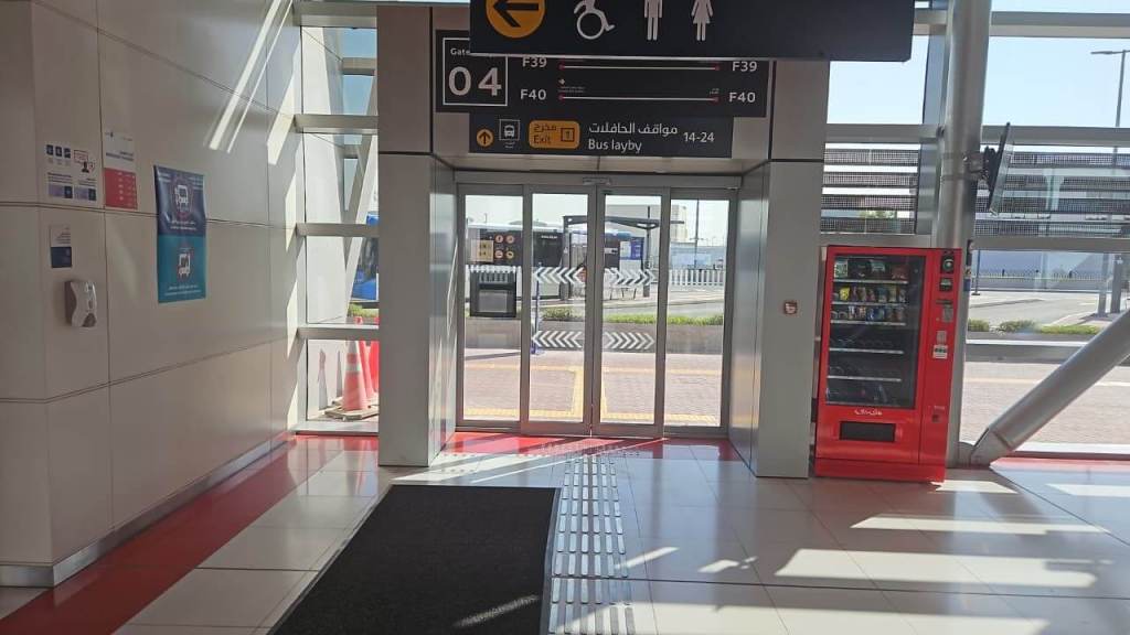 Interior view of a transit terminal featuring glass doors, directional signs for gates F39 and F40, and a red vending machine nearby. The area is well-lit with a clean floor and large windows allowing visibility outside.
