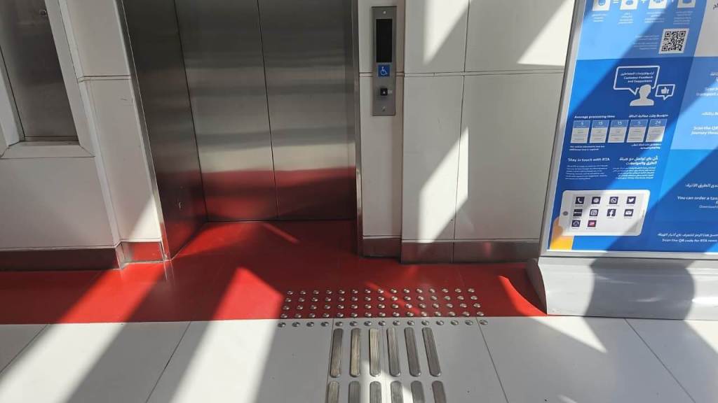 Elevator entrance with a wheelchair accessible button, adjacent to a sign with information and QR code, featuring a red and white floor design.