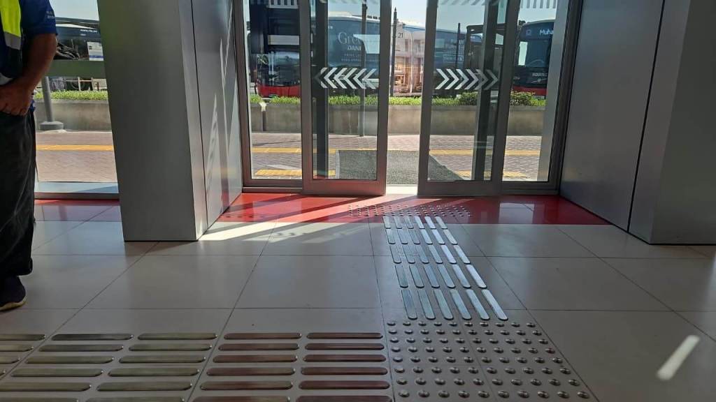 Interior view of a transit station showing automatic glass doors, tiled floor with tactile indicators, and a person standing nearby.