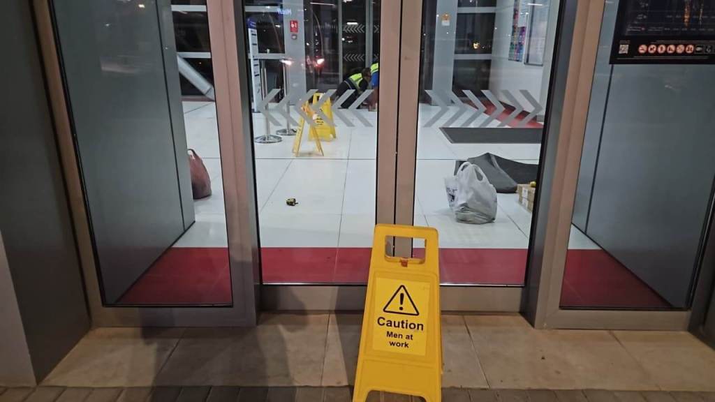 A caution sign reading 'Caution Men at work' placed in front of glass doors, indicating ongoing work inside the building, with reflection visible in the glass.