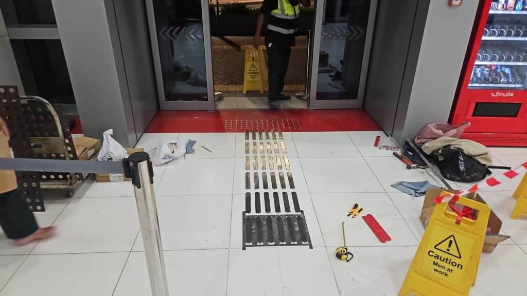 A worker in a safety vest stands near a partially completed floor entrance, with maintenance equipment and caution signs visible. Tools and materials are scattered on the floor, indicating ongoing work.