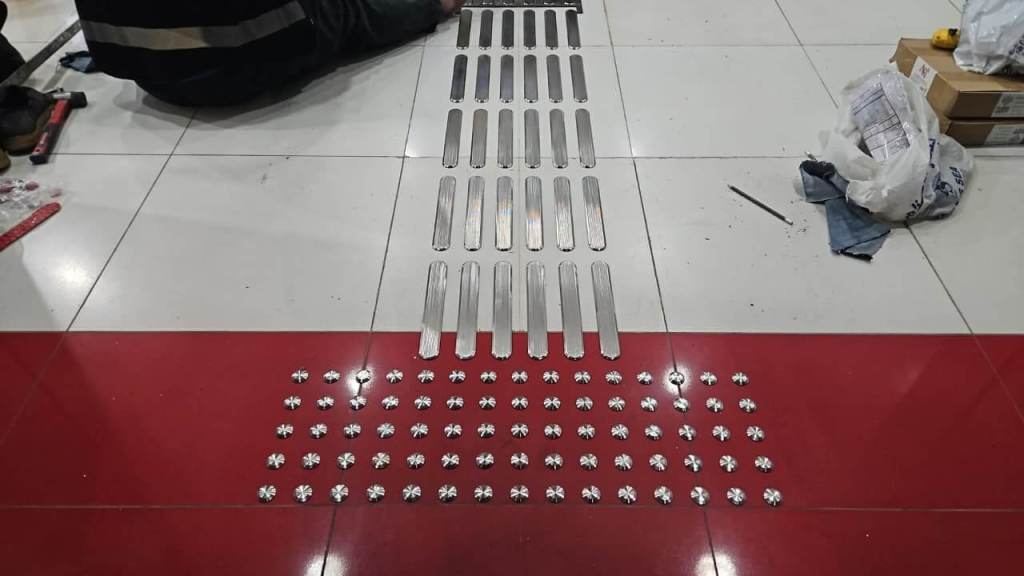 A series of metal strips and circular components arranged on a tiled floor, with a person's hand visible, preparing for installation.