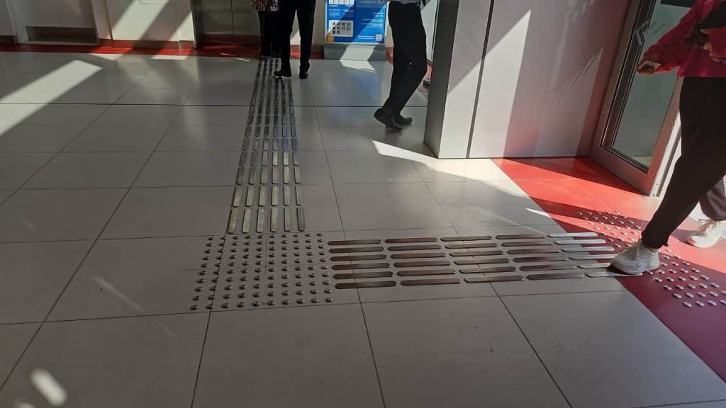 Interior view of a public space with tiled flooring, featuring tactile guiding strips and people walking in different directions.