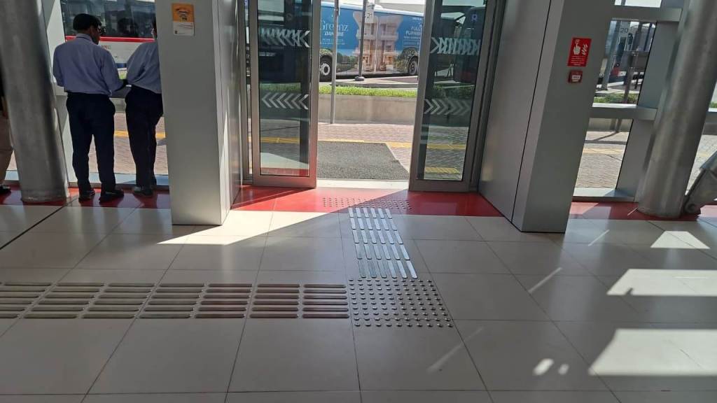 Interior view of a modern transit station featuring two people waiting near transparent sliding doors, with tiled flooring and visible bus traffic outside.