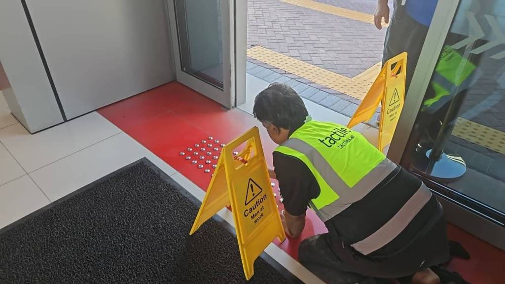 A worker in a reflective vest is installing tactile indicators on a red surface at a building entrance, with yellow caution signs placed nearby.