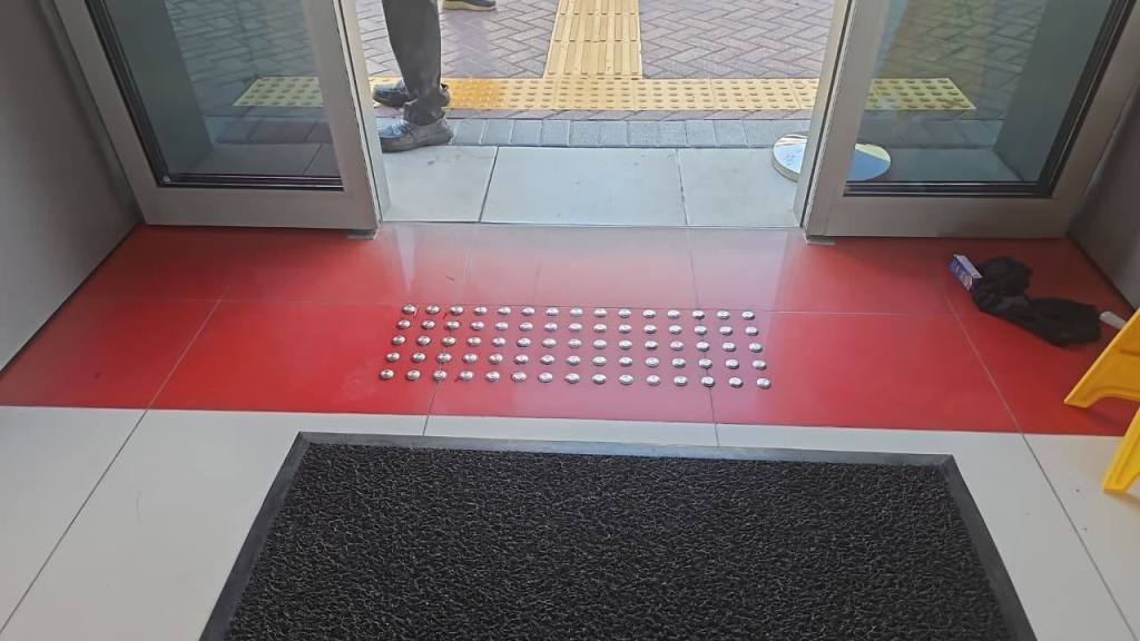 Entrance view showing red flooring with tactile paving dots and a welcome mat, leading to an outdoor area with yellow tactile indicators.