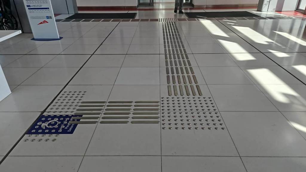 Interior floor of a transportation facility featuring tactile guidance indicators for visually impaired individuals, alongside a sign indicating accessible services.