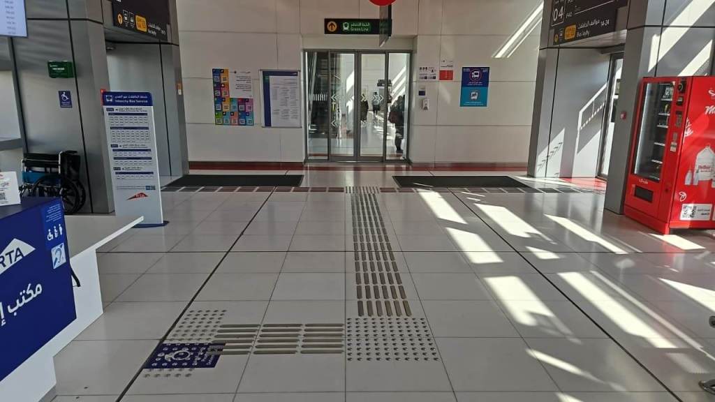 Interior view of a transit station featuring a ticket counter, elevators, and vending machines with directional signage and tactile paving on the floor.