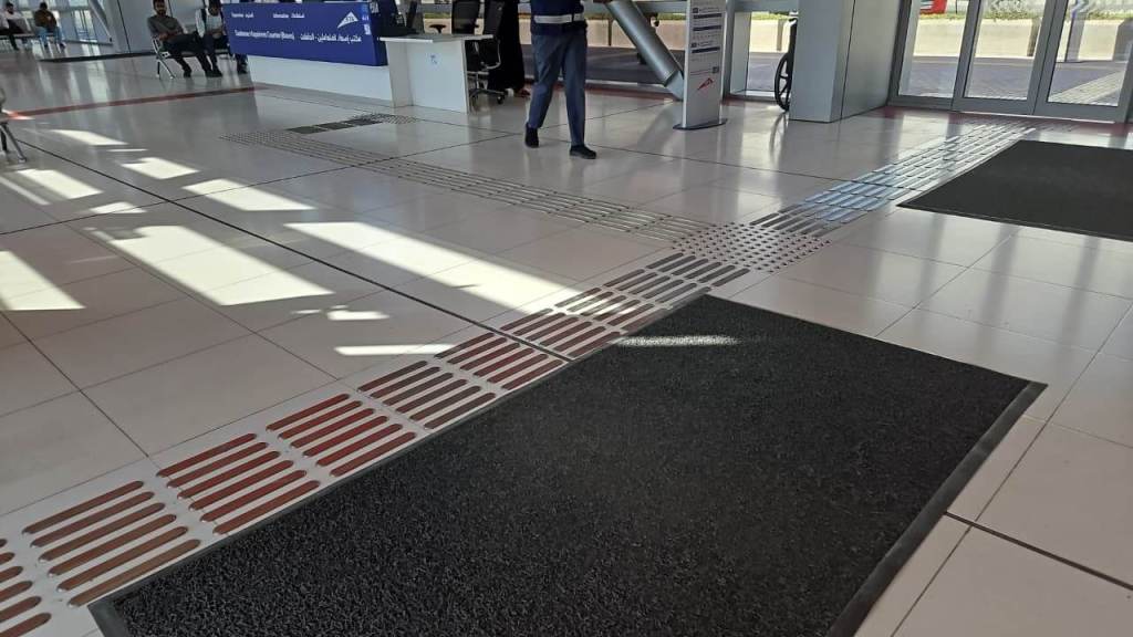 Interior view of a modern public building showing tiled flooring with textured strips and dark mats, sunlight casting shadows across the space.