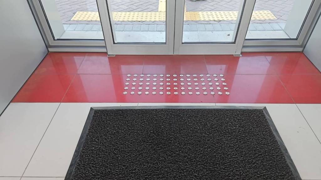 Interior entrance with a textured mat and a tiled floor featuring a red section and a row of circular tactile indicators leading to the entrance doors.
