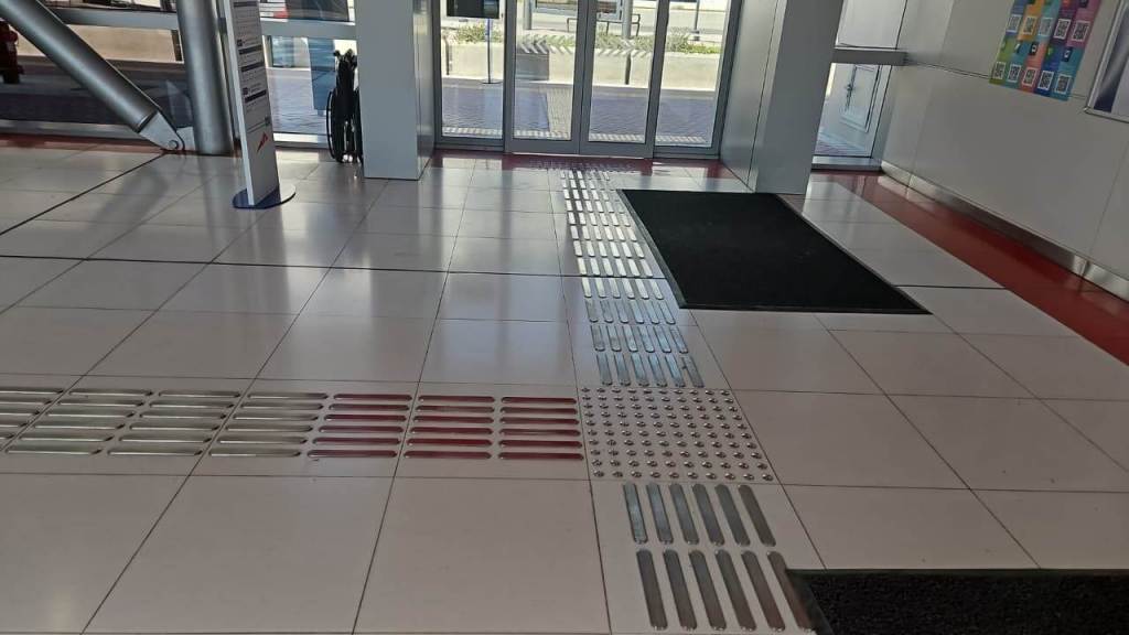 Interior view of a building entrance showcasing a tiled floor with tactile guide strips and mats, leading toward a glass door.