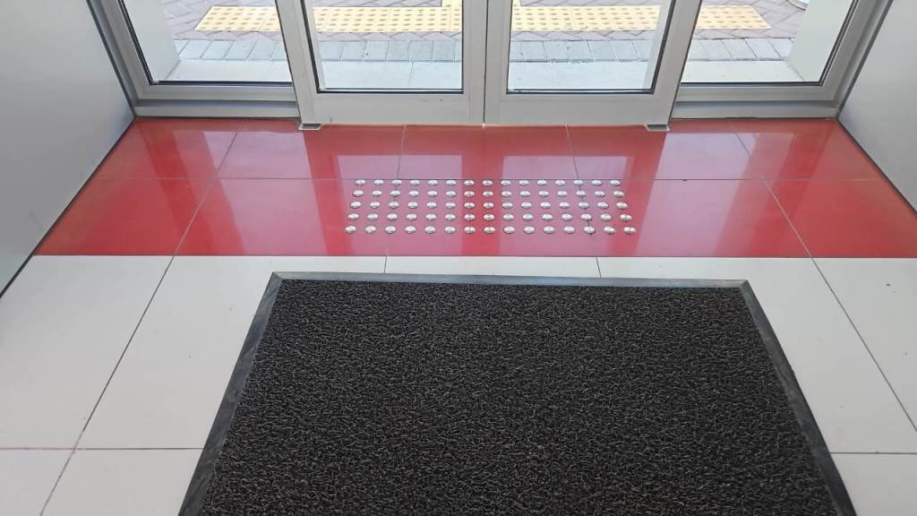 Entrance area with red and white tiled floor, featuring a textured mat and tactile paving for navigation.
