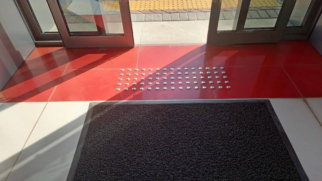 Interior view of a doorway featuring glass doors and a red tiled entry with tactile indicators. A black doormat is visible on the floor.