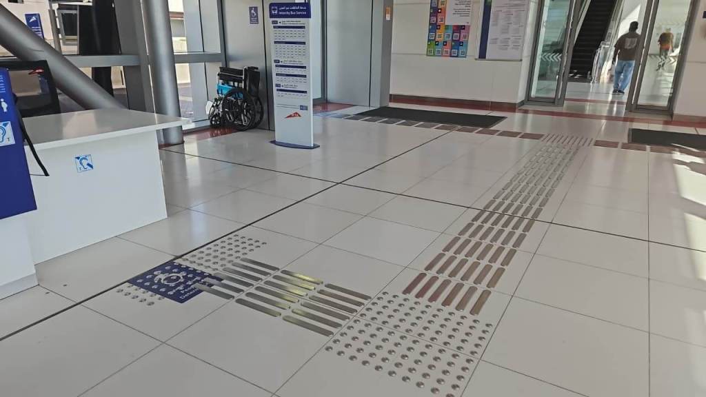 Interior of a public building showcasing a spacious lobby area with tactile paving for accessibility, a wheelchair in the background, and informational signage.