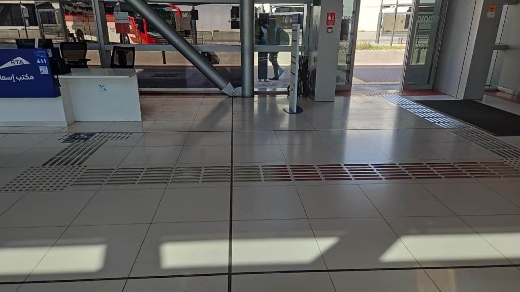 Interior of a transportation hub showing a ticket counter, tiled flooring with tactile paving, and large glass entrance doors.