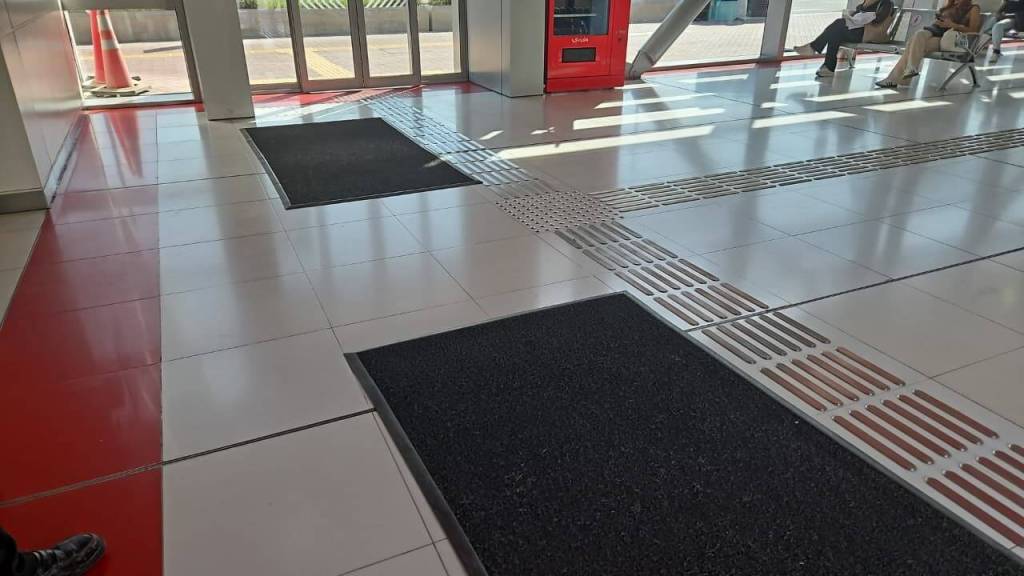 Interior view of a waiting area with a tiled floor, featuring mats in two locations, large windows allowing natural light, and a red vending machine in the background.