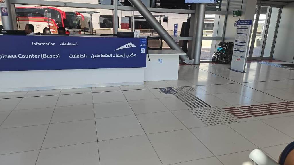 Information desk for bus services with a sign in English and Arabic. The area is brightly lit and features tiled flooring with accessibility markings, including a wheelchair symbol nearby.