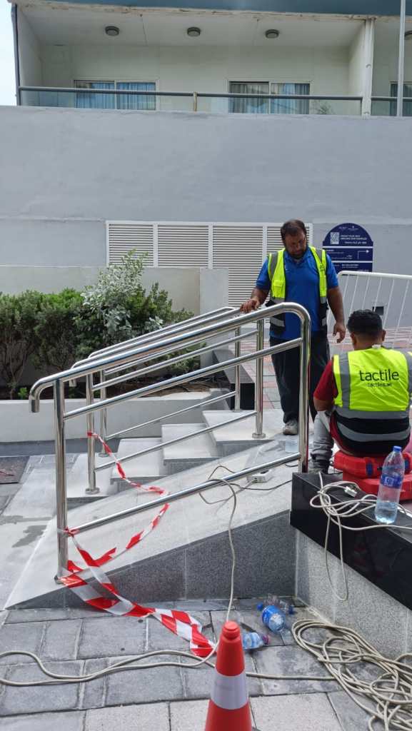 Two workers in safety vests are inspecting a ramp with handrails under construction. Construction materials and tools are visible, along with a traffic cone and caution tape.