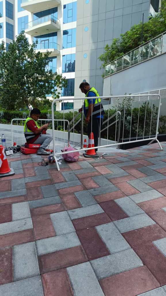Two construction workers in safety vests working on a temporary barrier near a building. One worker is sitting on the ground with tools, while the other is standing and adjusting the barrier. The area features patterned paving stones and traffic cones.