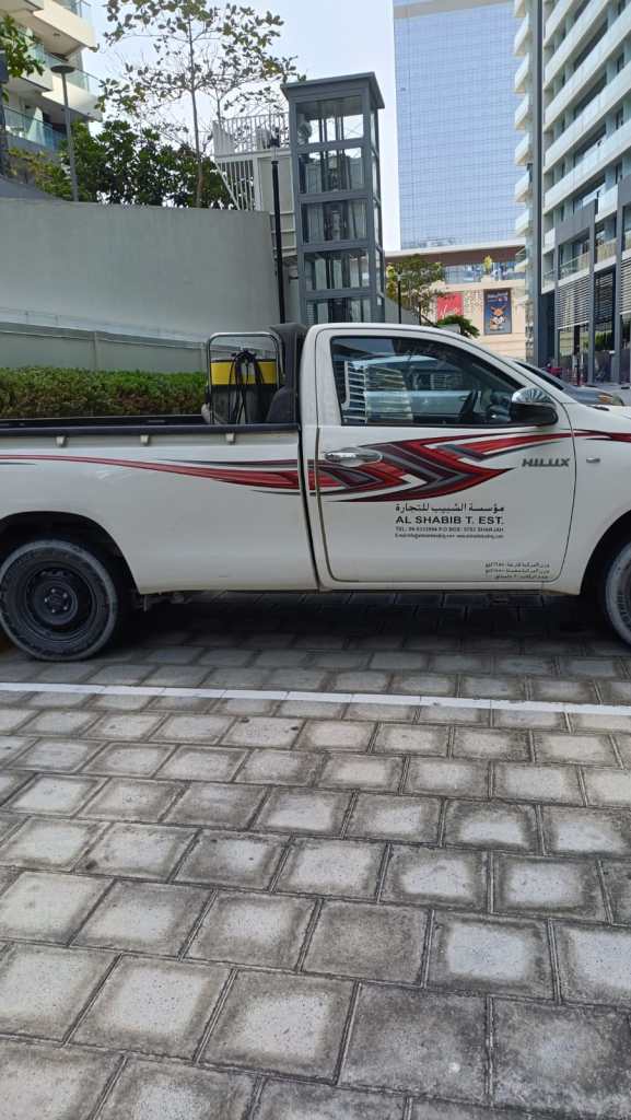 A white pickup truck parked on a paved surface, featuring a red and black design on the side and displaying the text 'AL SHABIB T. EST' on the door.