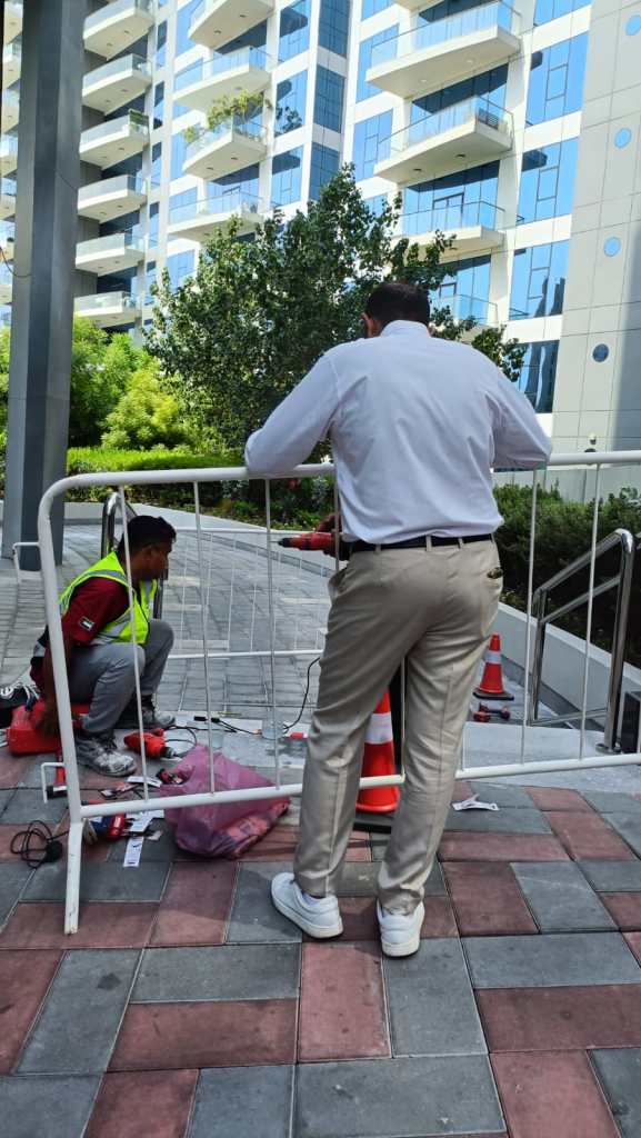 A man in a white shirt supervises a construction site while another worker in a neon vest is seated on the ground, surrounded by tools and traffic cones, near a modern building.