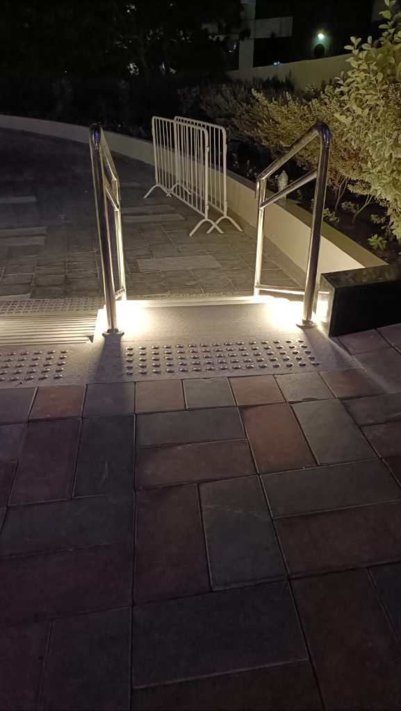 A well-lit set of stairs with stainless steel handrails, leading down to a paved area at night, with a protective barrier in the background.