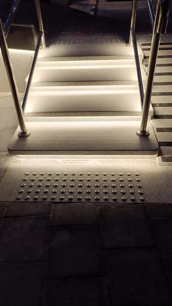 Illuminated outdoor stairs with handrails, featuring textured warning strips at the base for safety.