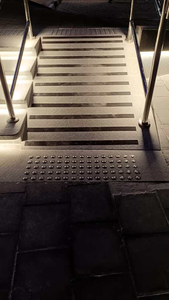 Close-up view of illuminated stairs with tactile paving for accessibility, featuring contrasting strip patterns and handrails.
