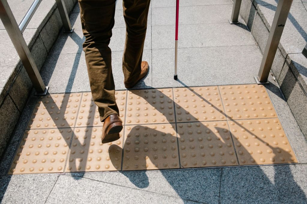 A person walking with a white cane on textured tactile paving, casting a shadow on the ground.
